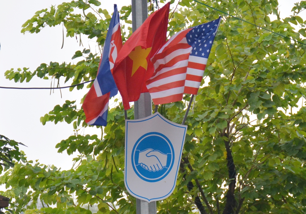 Bên dưới cờ 3 nước là biểu tượng hai bàn tay bắt chặt, thể hiện tinh thần và khát vọng của nhân dân yêu chuộng hòa bình thế giới trước cuộc gặp giữa hai nhà lãnh đạo Mỹ và Triều Tiên.